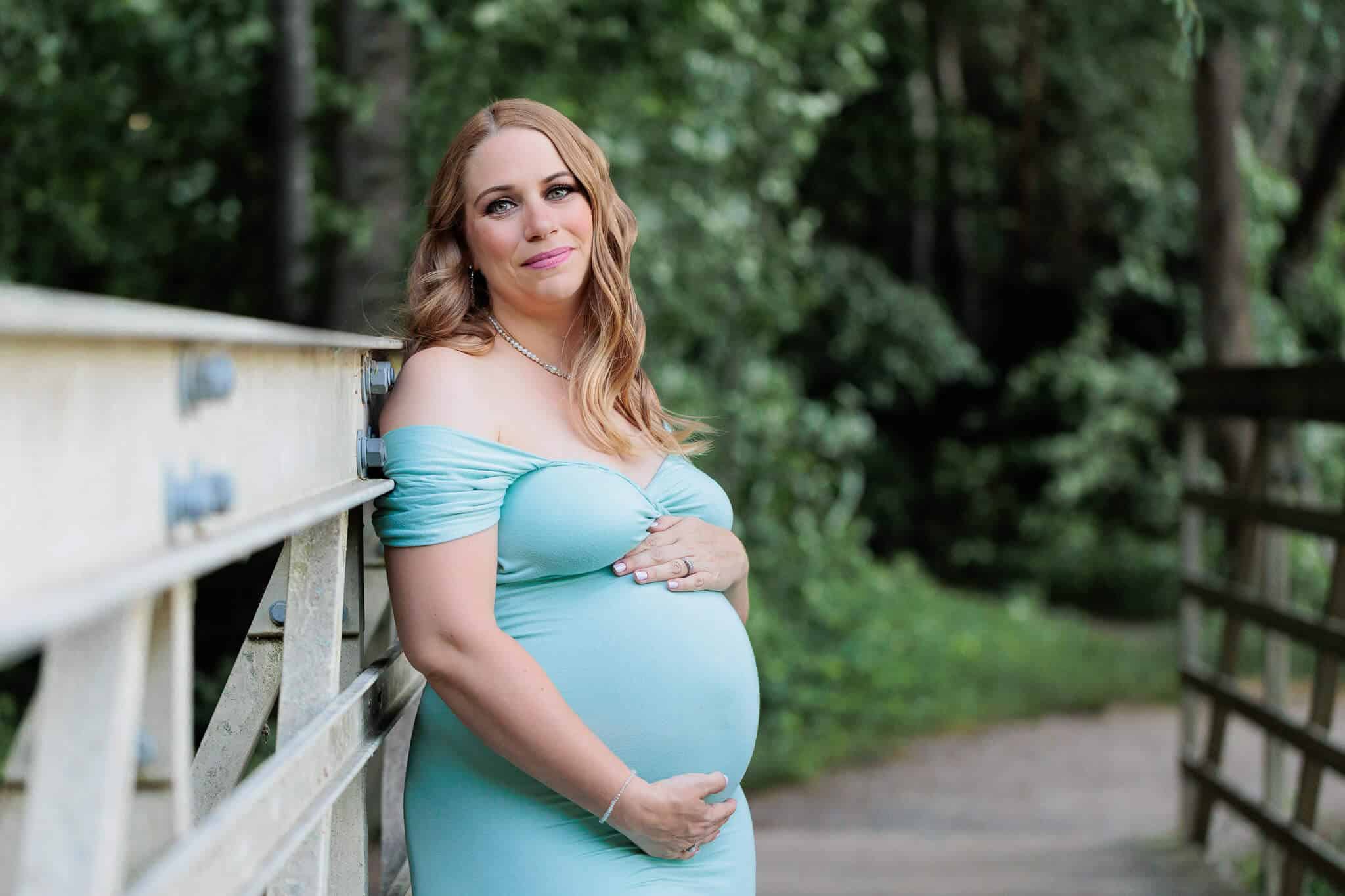 A woman hugging her pregnant belly in a blue dress and leaning up against a bridge being featured on a blog about Gainesville VA midwives.