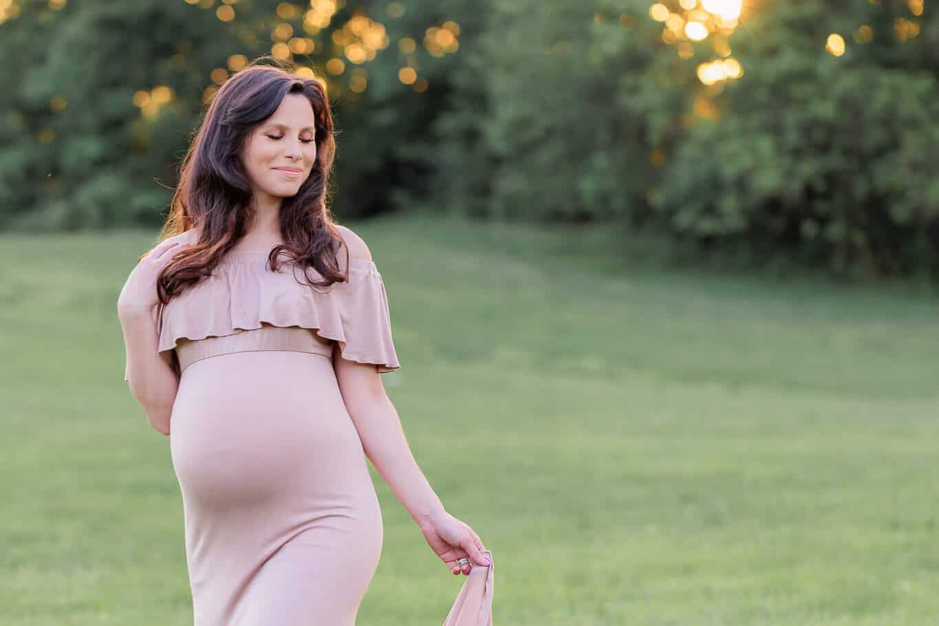 A beautiful pregnant woman posing in a tan dress with the sun setting behind the trees, featured on a blog about Northern Virginia OB/GYN practices.
