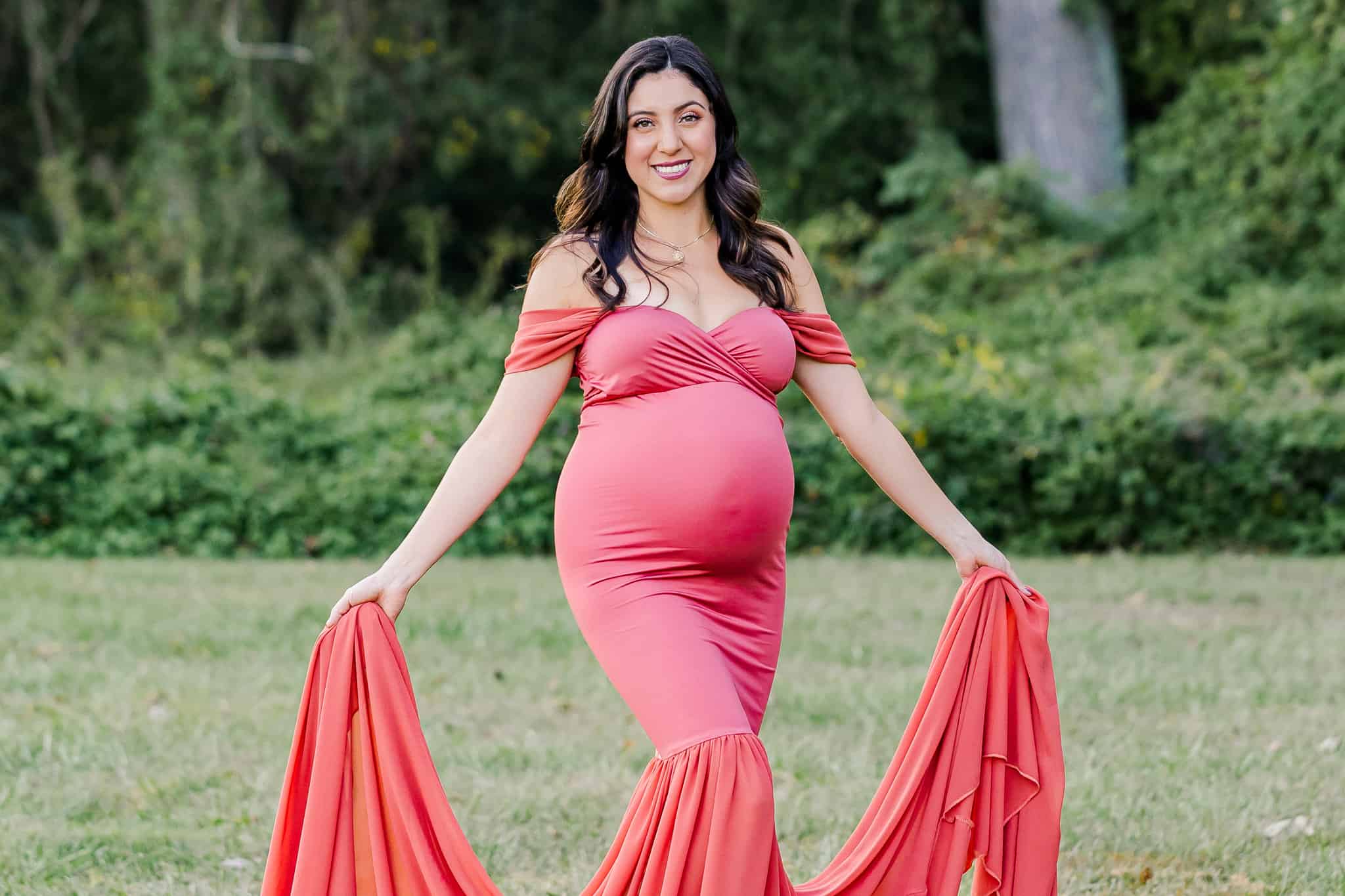 A pregnant woman standing in a field and holding the train of her rose colored dress being featured in a blog about baby shower venues Northern Va.