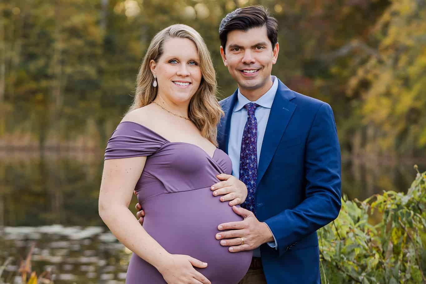 A pregnant woman in a purple dress posing with her husband in front of a pond, being featured on a blog about Babycito.