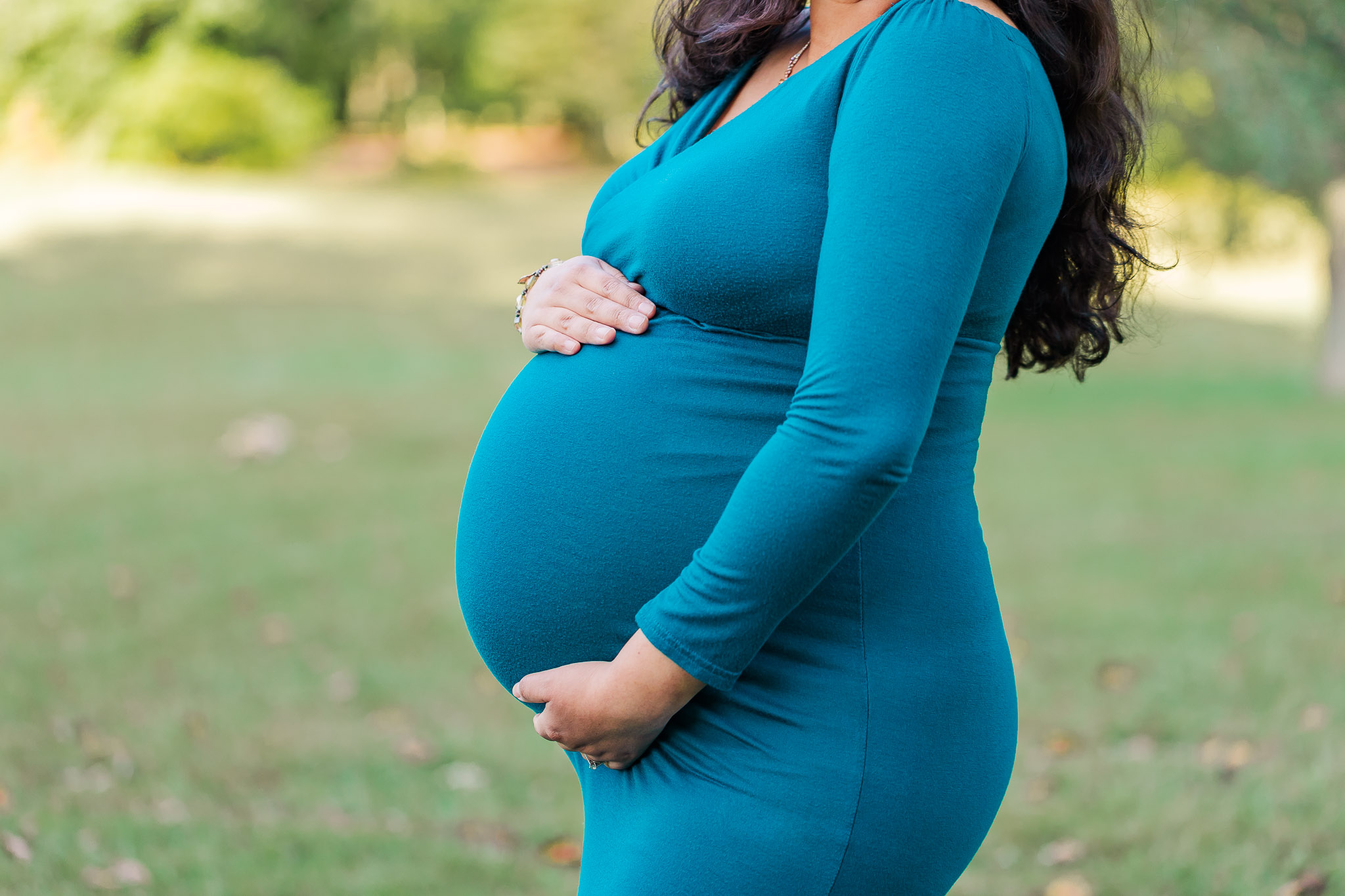 A closeup of a mother-to-be cradling her belly in a field and wearing a dark teal dress, being featured on a blog about Birth in Bloom.