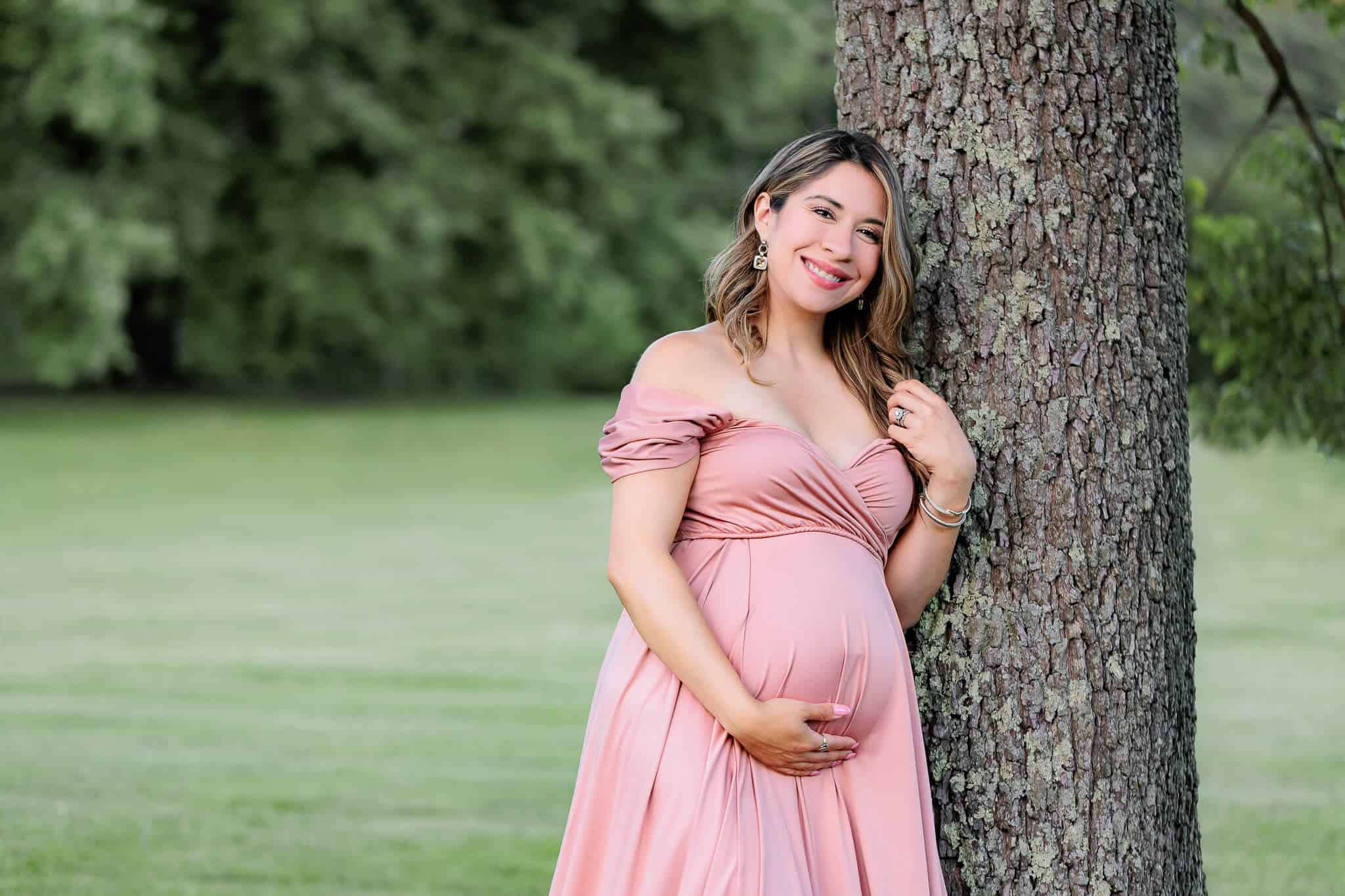 A beautiful pregnant woman in a pink dress holding her belly and leaning against a tree at a park, being featured on a blog about Inova Alexandria Birthing Center.