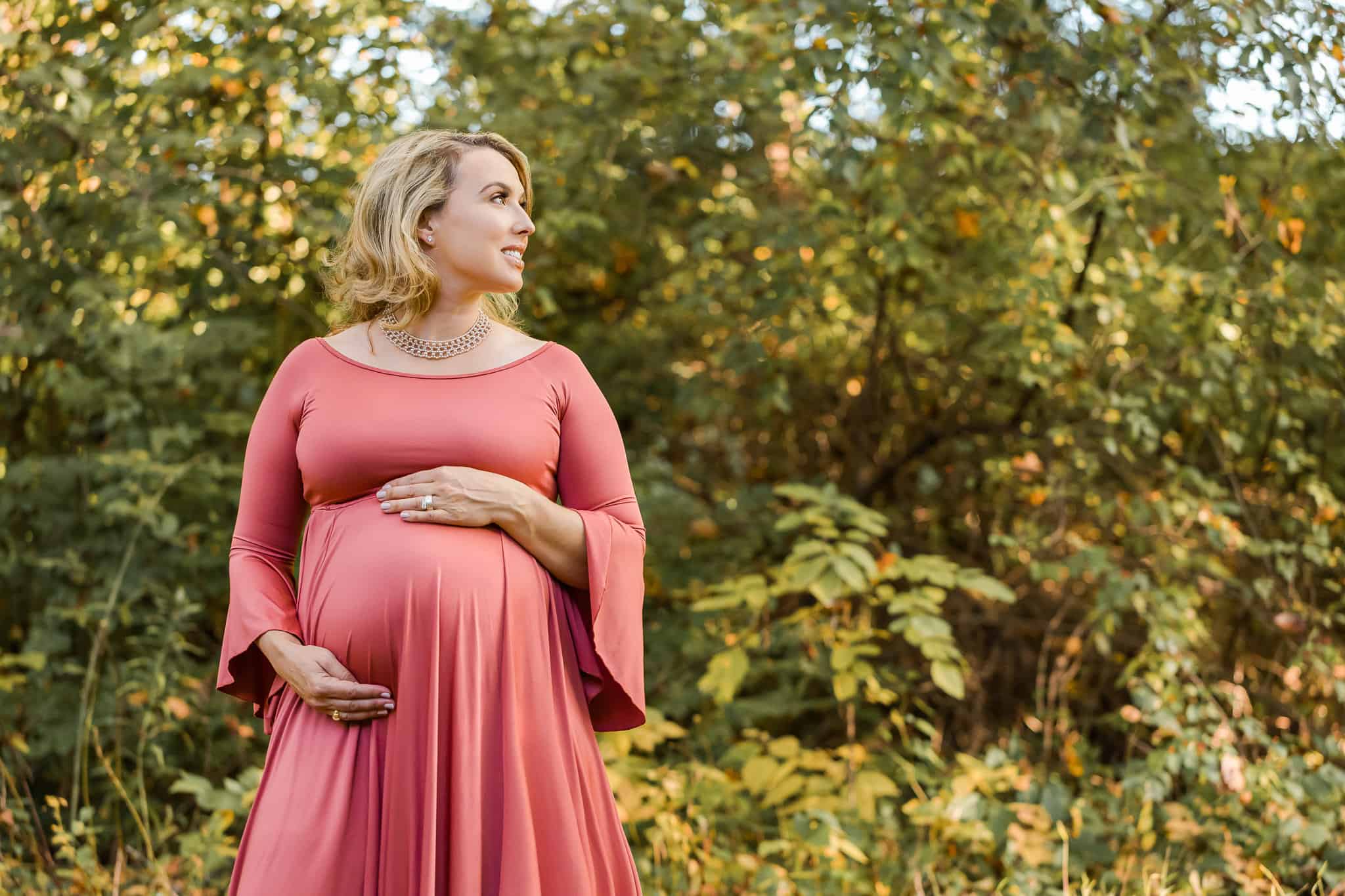 A beautiful mother-to-be in a rose colored dress posing in front of some fall foliage, featured on a blog about placenta encapsulation Northern Virginia.