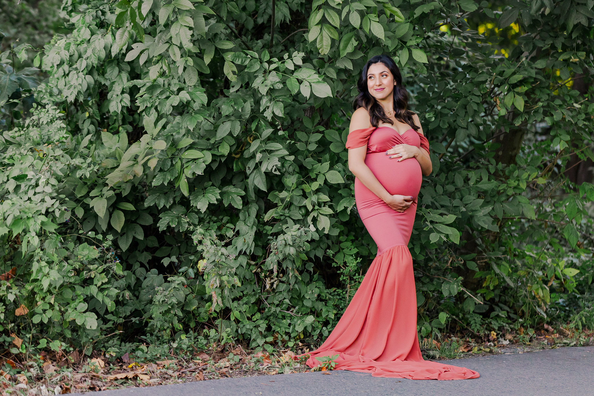 A beautiful pregnant woman in a pink dress standing in front of green bushes and hugging her belly, being featured on a blog about prenatal massage Alexandria.