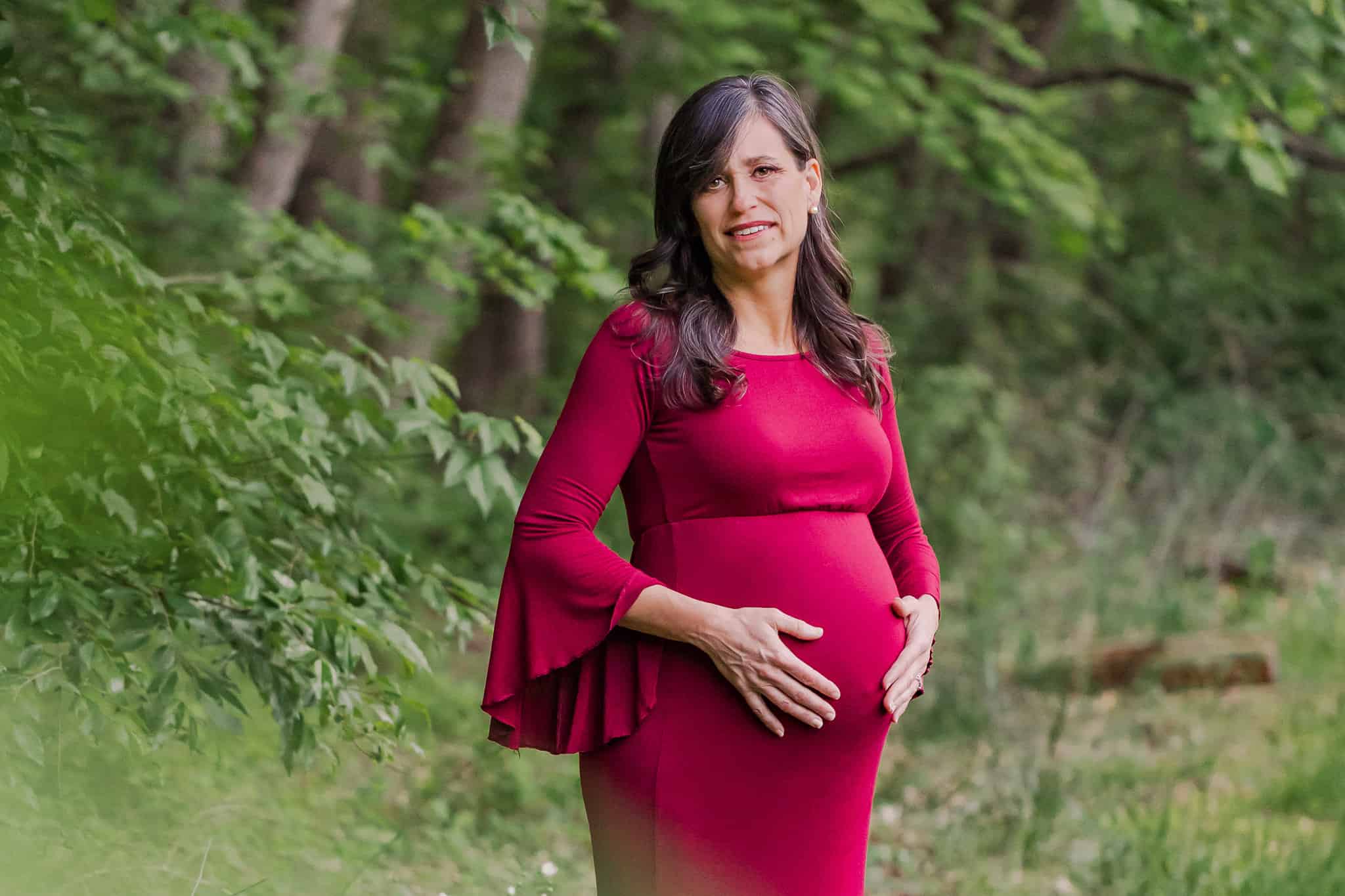 A pregnant woman in a red dress holding her belly and posing in the woods, being featured on a blog about Sibley Hospital maternity.