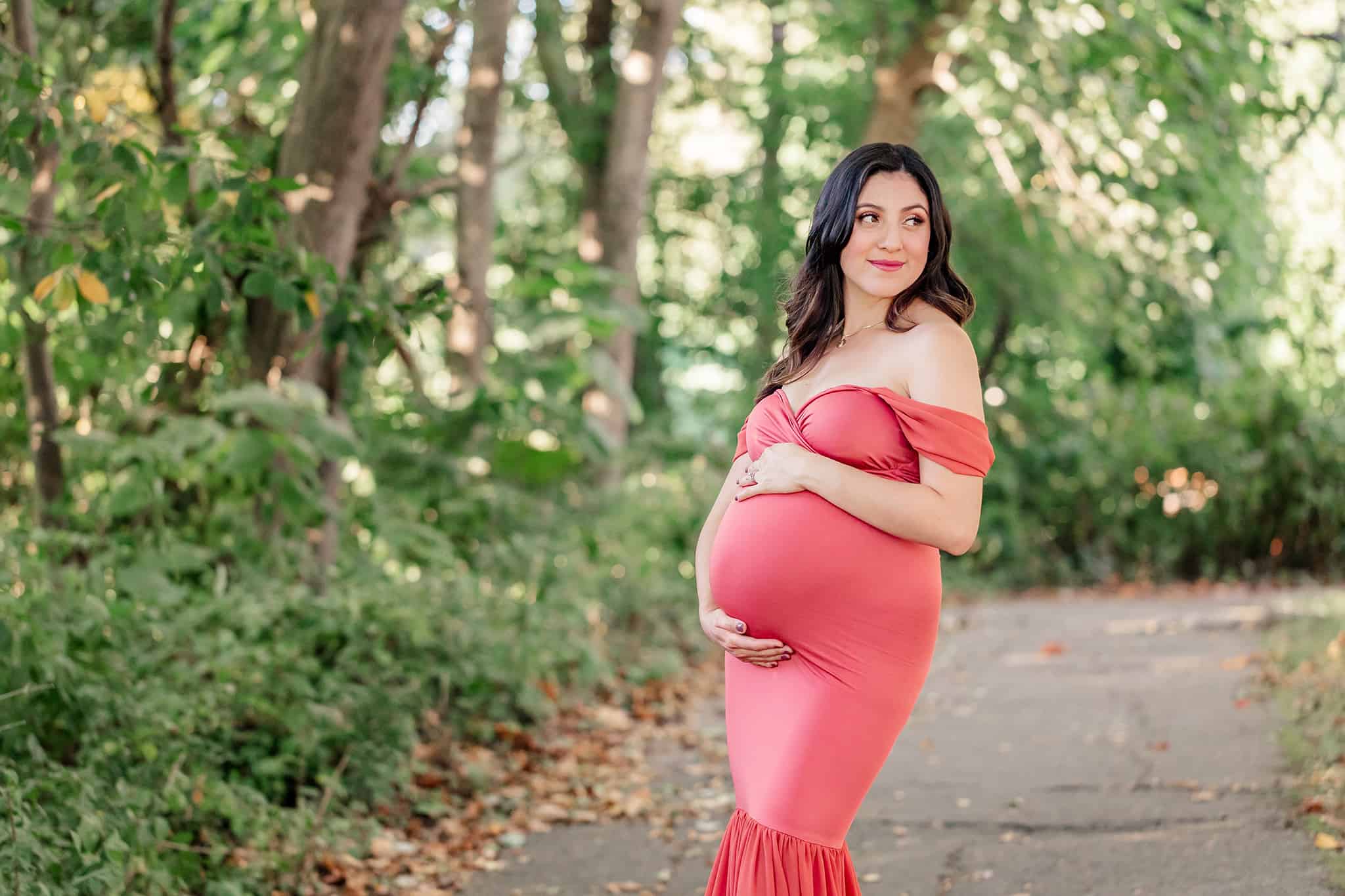 A blog about Physicians and Midwives Alexandria featuring a photo of a pregnant woman in a pink dress posing on a trail in the woods and holding her belly.