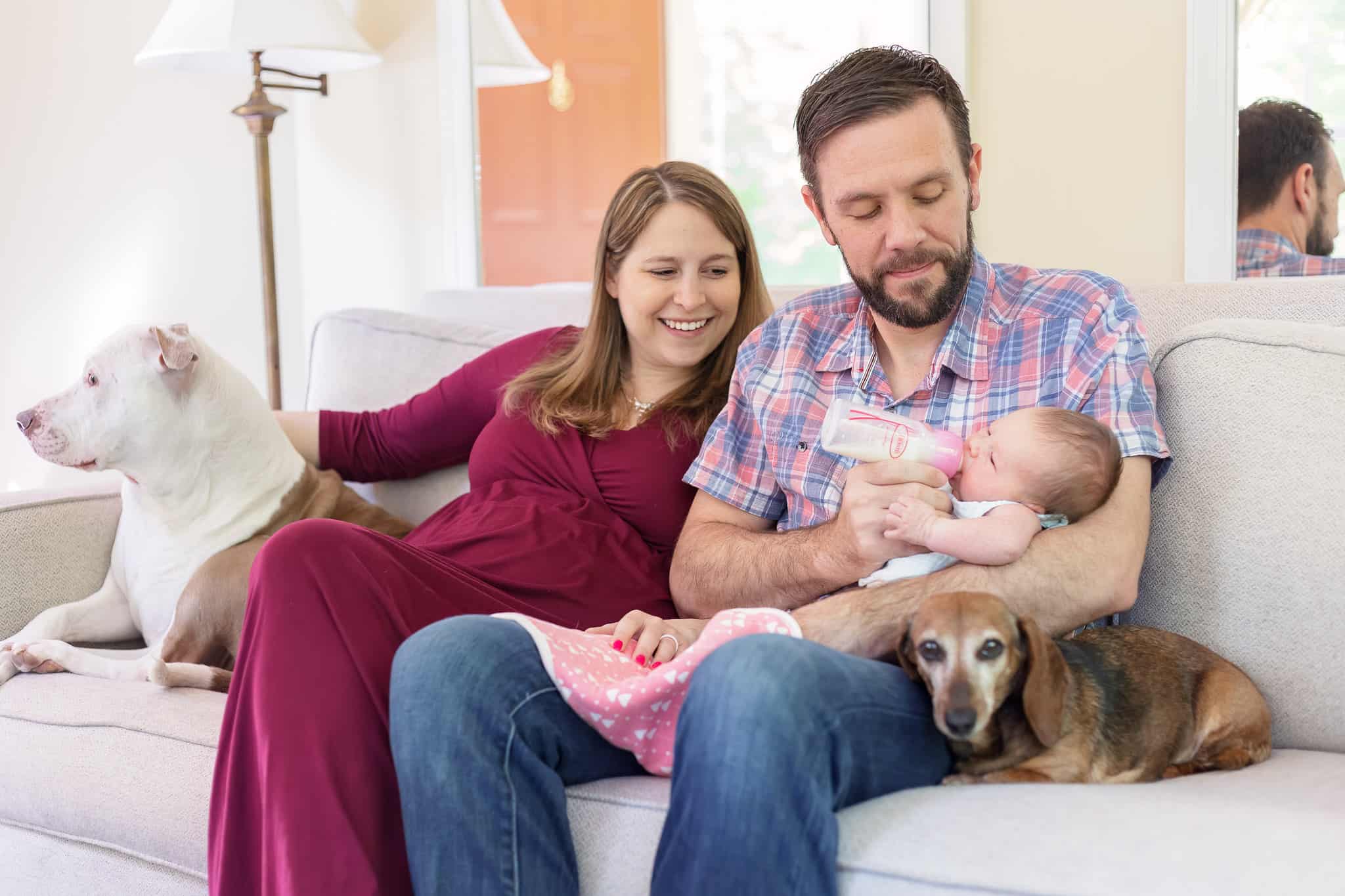 A mom and dad sitting on the couch in their Manassas VA home feeding their newborn baby and snuggling with their two dogs.