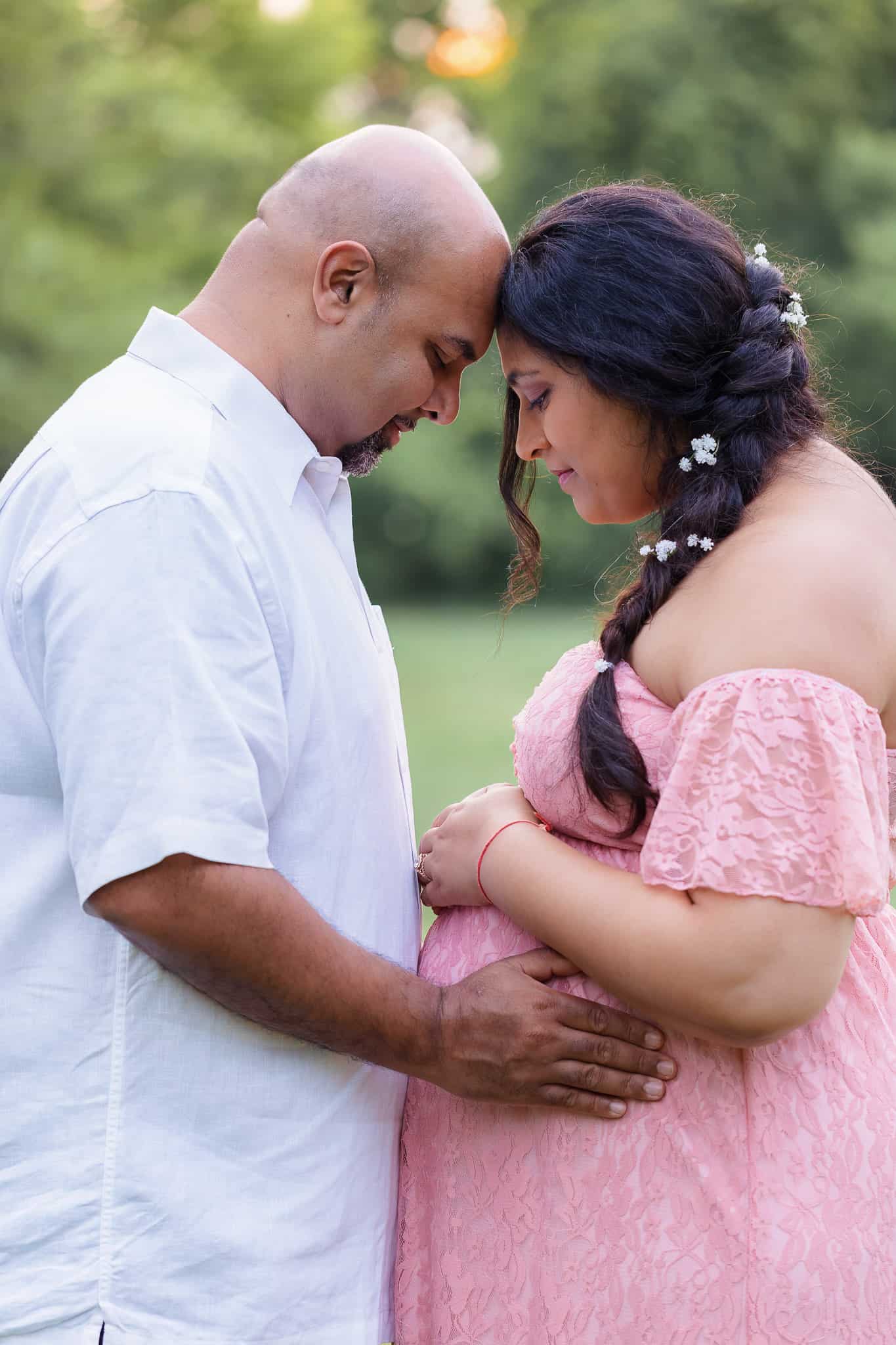 An expecting mom and dad touching foreheads and embracing the pregnant belly in a park at sunset.