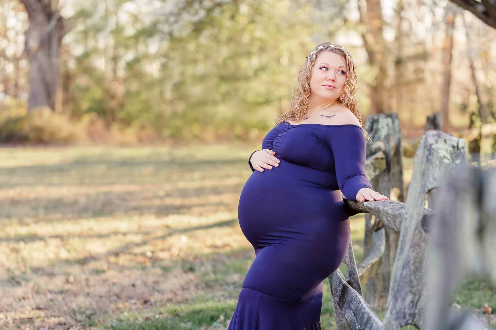 A pregnant mom in a blue dress leaning against a fence at Burke Lake Park.
