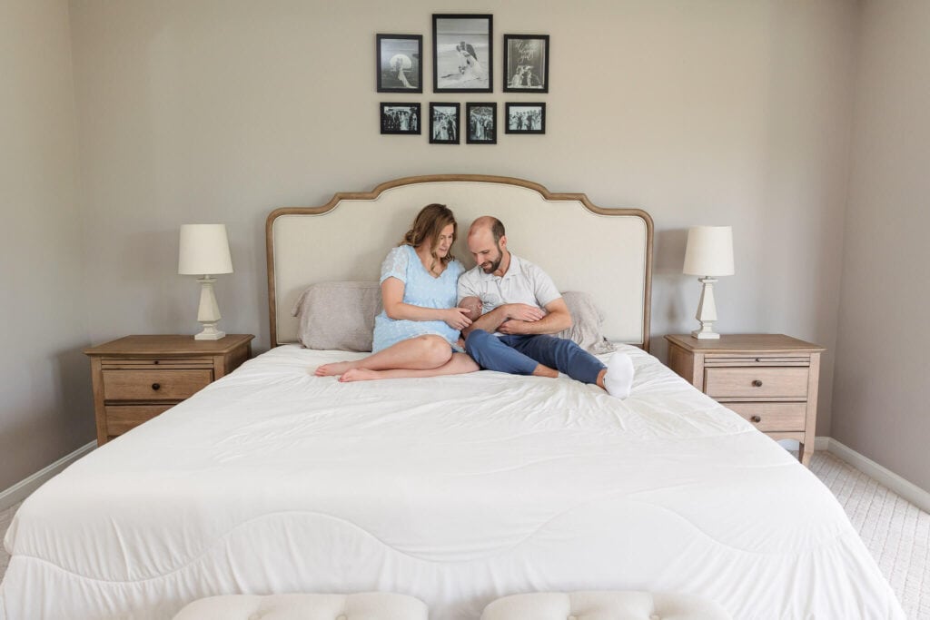 A mom and dad sitting on their bed snuggling their baby boy during a lifestyle newborn session in their home.