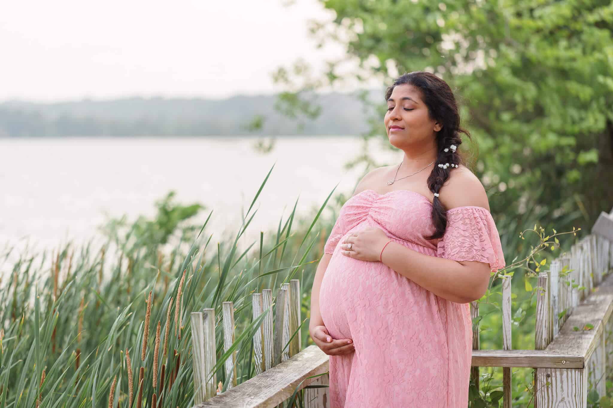 A pregnant mom holding her belly and closing her eyes as she feels the breeze coming off the river in front of her at Jones Point Park, one of my favorite photoshoot locations in Northern Virginia.