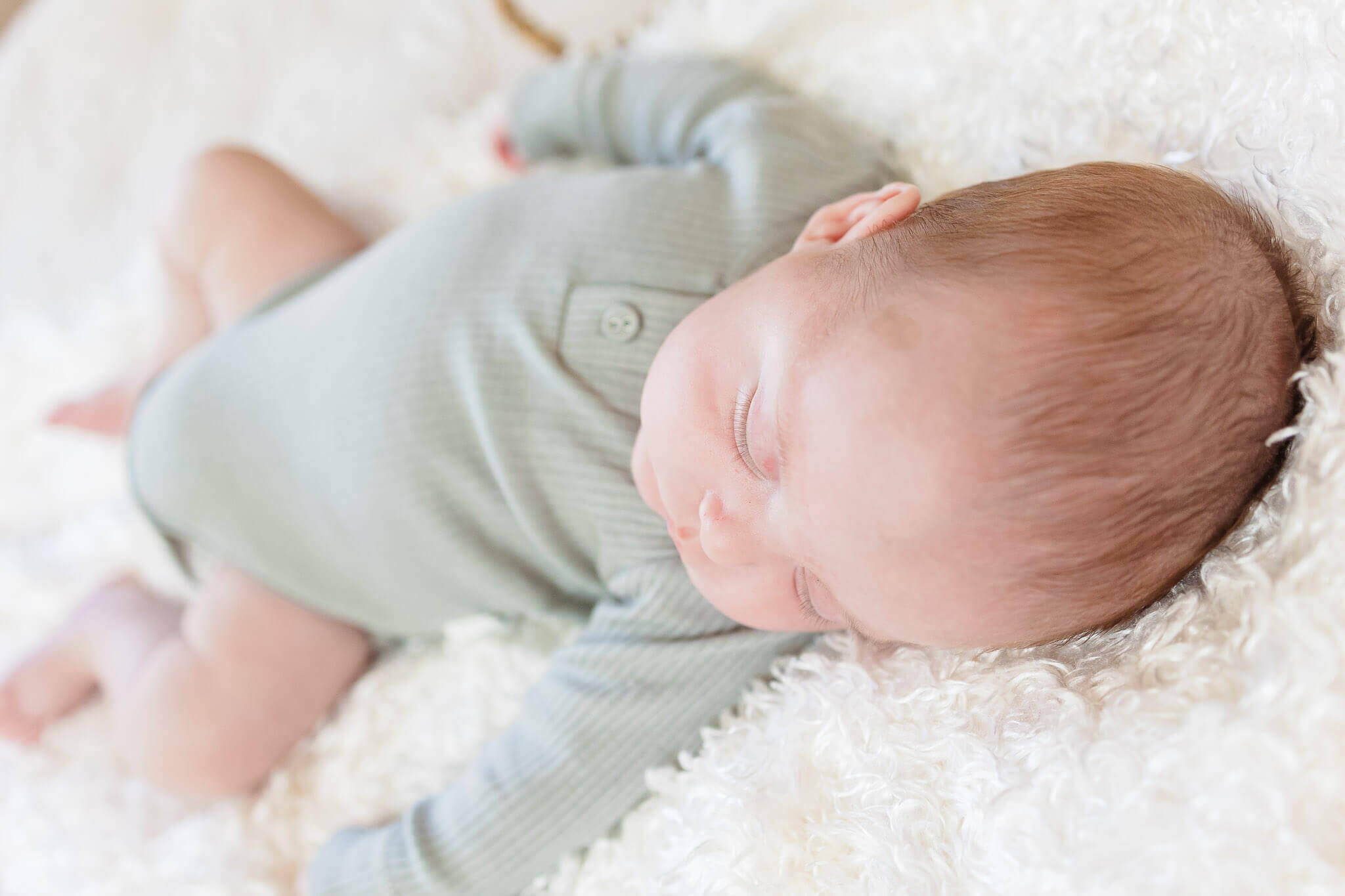 A newborn boy wearing a green onesie and sleeping on a cream blanket.