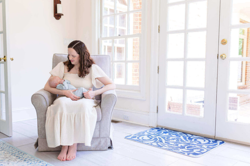 A mom holding her newborn son and sitting on a chair in their home.