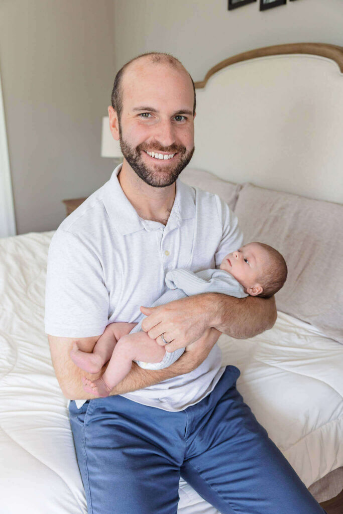 A dad holding his newborn son and sitting on the edge of the bed