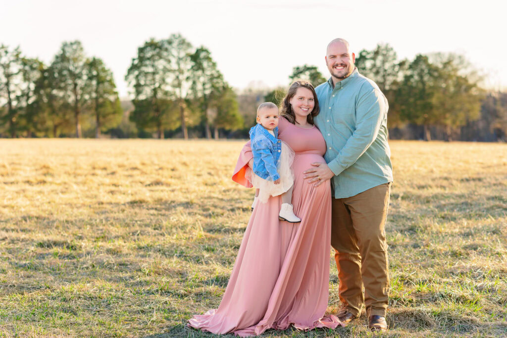 Expecting mom in a dusty pink maternity gown with her husband in a sage button-up and their toddler in a denim jacket